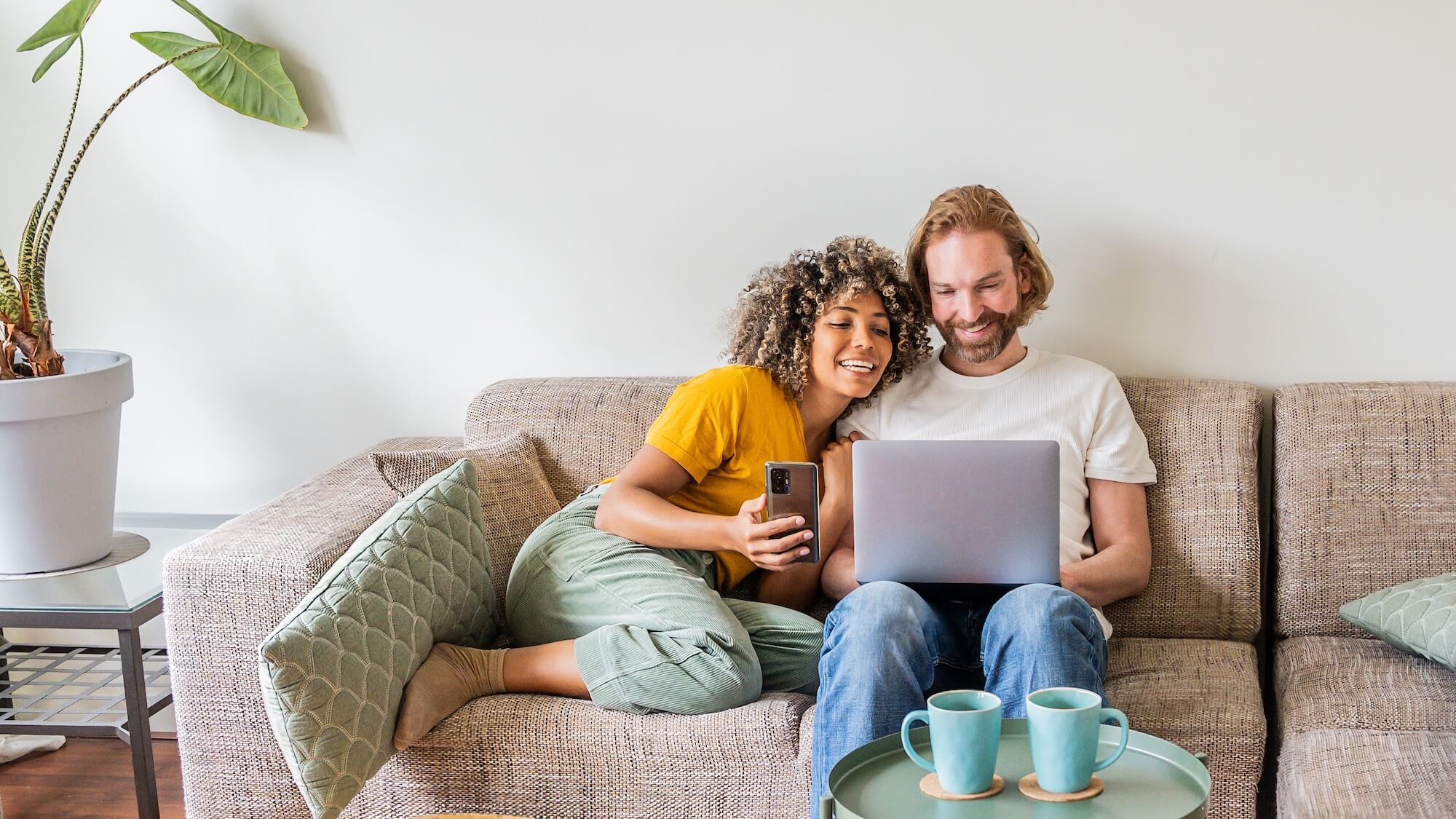 Two people sitting on a modular sectional couch with a laptop and coffee mugs, surrounded by plants and a plant stand.